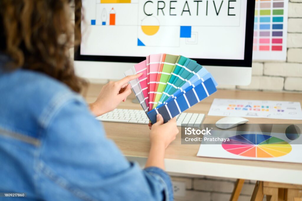 Femme choisissant des couleurs à utiliser pour son projet créatif sur un ordinateur de bureau à l'aide d'échantillons de couleur sur carton et d'un cercle chromatique.