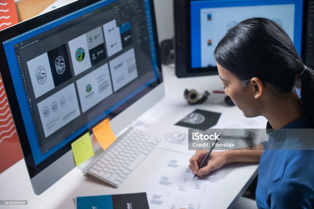 Jeune femme tenant traçant des lignes au crayon sur des images imprimées et en étant assise en face de ses écrans d'ordinateur de bureau sur lesquels des images de logos figurent.